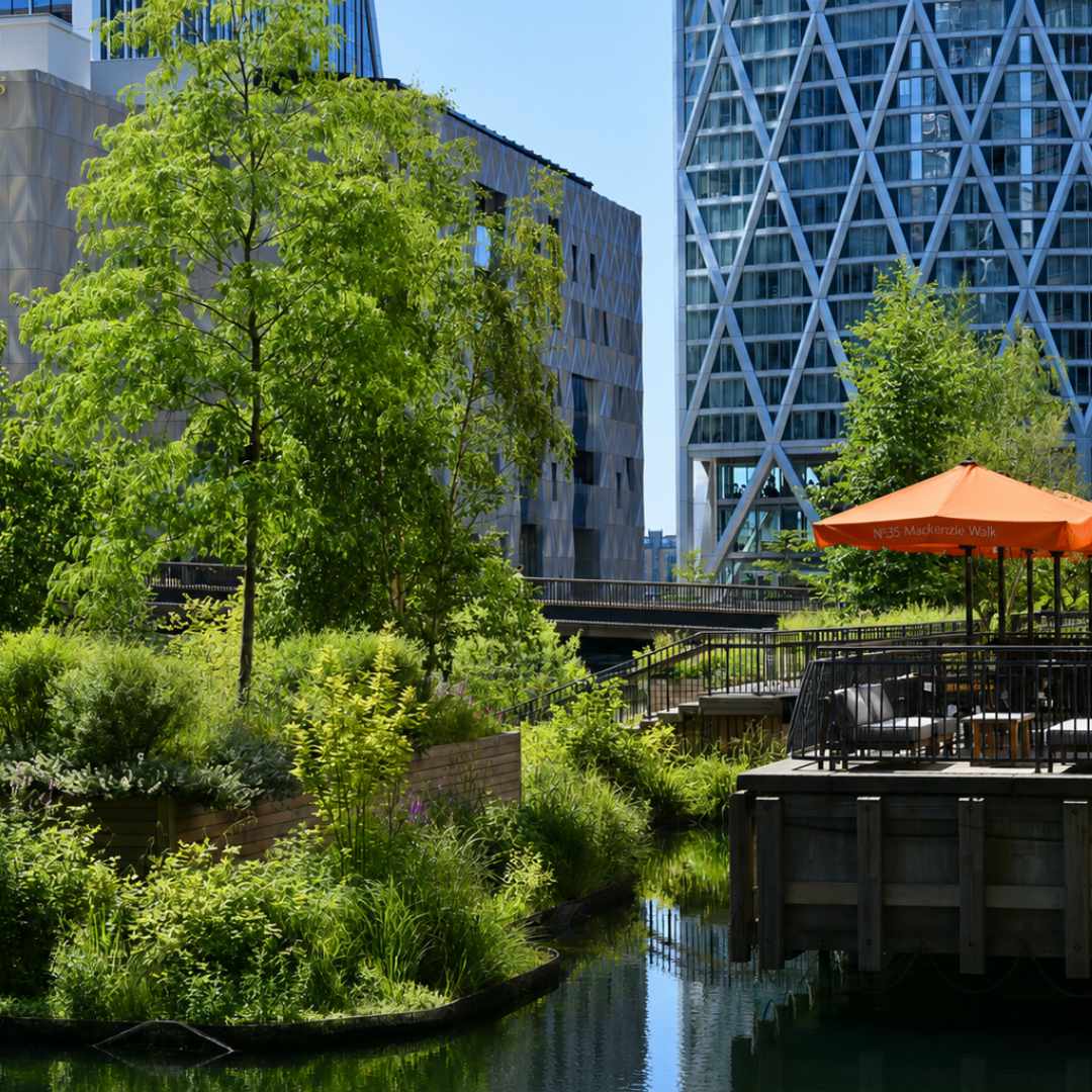 Landscaped canal garden with lush planting and an empty restaurant terrace beneath orange umbrellas, surrounded by modern Canary Wharf towers